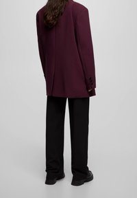 Vinröd kavaj med markerade axlar och knäppta ärmslut, kombinerad med svarta, löst sittande byxor och robusta skor. Slät textur överlag.