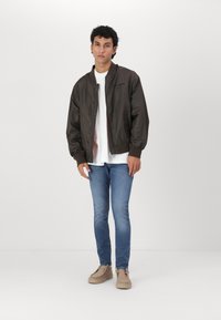 Brun bomberjakke med en glat overflade, hvid t-shirt underneath, blå jeans og beige sko. Modellen står foran en ensfarvet baggrund.