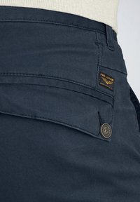 Pantaloni cargo blu navy con finitura testurizzata, caratterizzati da una tasca con un bottone metallico e un'etichetta con il marchio sulla vita.