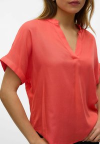 Blusa corallo a maniche corte con scollatura a V, realizzata in tessuto leggero. Presenta risvolti sulle maniche e una vestibilità rilassata. Tessuto morbido.