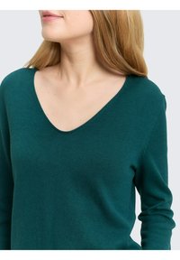 Türkisfarbener Strickpullover mit V-Ausschnitt und langen Ärmeln. Weiche Textur mit einer figurbetonten Passform, die ein glattes Finish und dezente Nähte zeigt.