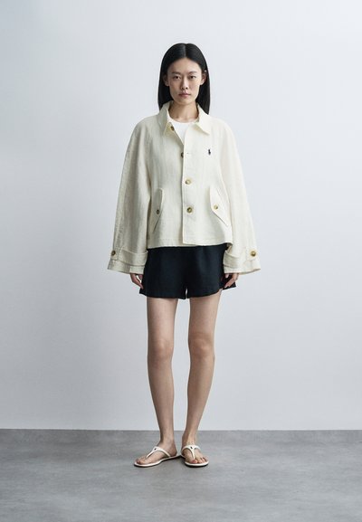 Žena nosiaca nadmernú krémovú bundu s vreckami na gombíky, čierne šortky a biele sandále, stojaca pred jednoliatym svetlosivým pozadím.