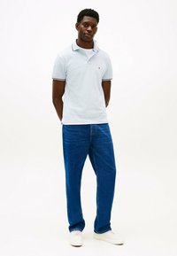 Camisa polo azul-claro com colarinho e detalhes em azul-marinho, combinada com jeans retos azul-escuro e ténis brancos. Roupa simples e casual.