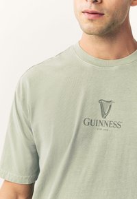 Svetlo zelena kratka majica z rokavi iz mehkega blaga, z zatemnjenim sivim logotipom Guinness in grafiko harfe, postavljenima v sredino prsnega koša.