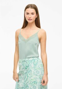 Camiseta de tirantes ligera de color verde claro con detalles de encaje. Combinada con una falda de diseño en tonos similares de verde y azul. Textura suave.