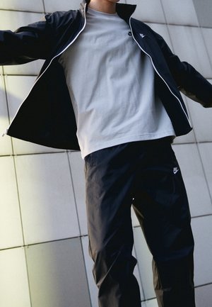Personne portant une veste noire Nike, un t-shirt gris et un pantalon noir Nike, debout les bras écartés devant un mur carrelé.