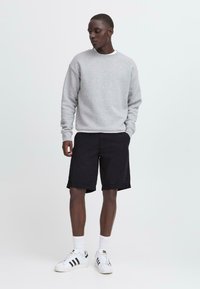 Man som står med händerna i fickorna, iförd en ljusgrå sweatshirt, svarta shorts, vita strumpor och vita sneakers med svarta ränder.