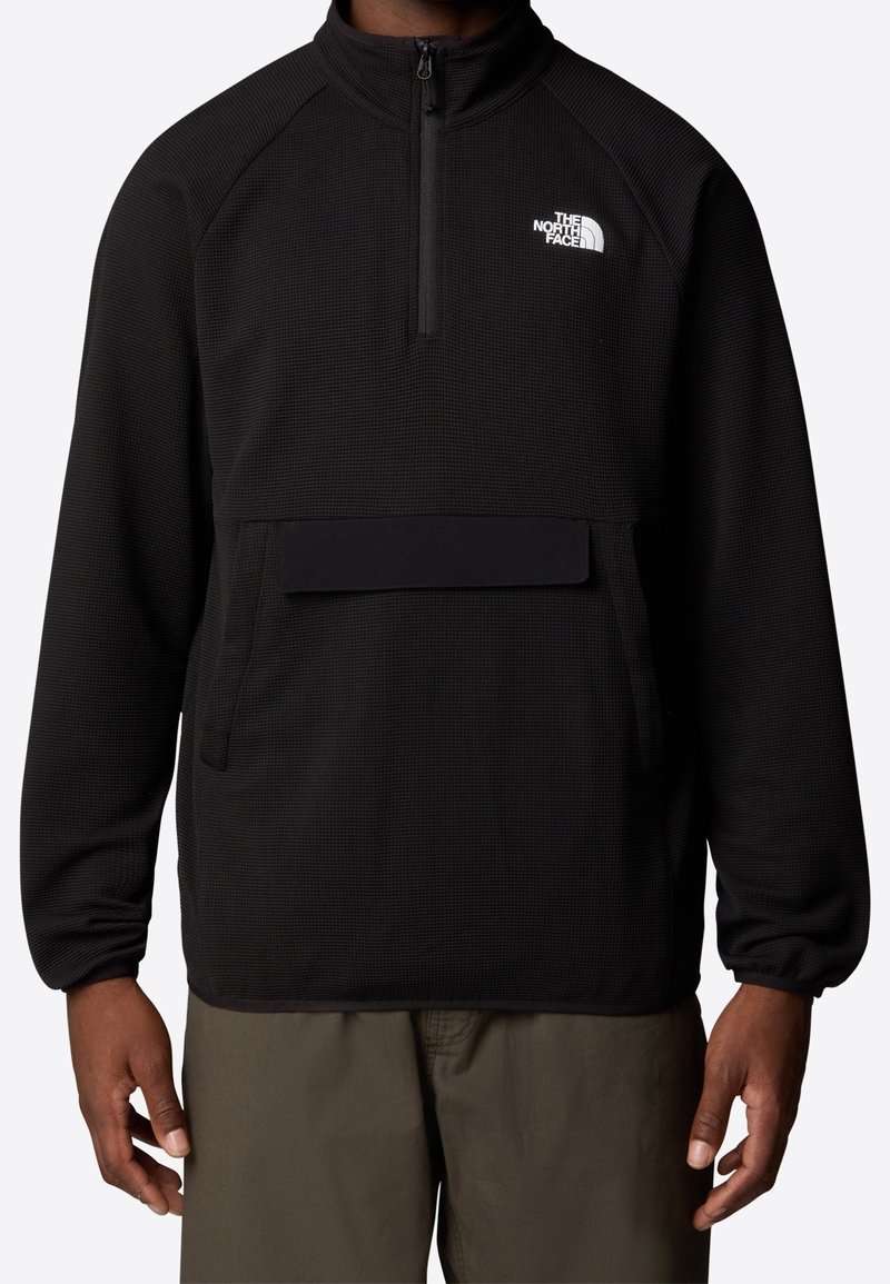 Pull noir à manches longues avec une fermeture éclair partielle, poche avant et logo blanc "The North Face" sur la poitrine, porté avec un pantalon vert olive.