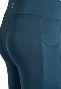 Mörk teal sportleggings med sömlös design, utrustade med en liten logotyp nära midjebandet och en diskret sidoficka för förvaring.