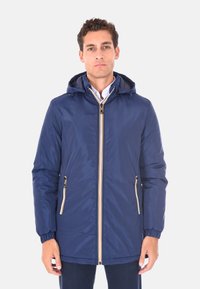 Uomo che indossa una giacca a capo con cappuccio di colore blu navy, con tasche con zip e cerniera frontale, sopra una camicia bianca con colletto.