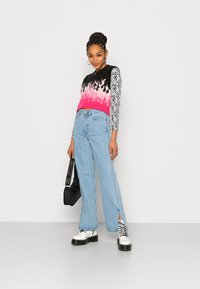 Sweater de malha com padrão de chama em preto, rosa e branco, com mangas longas em padrão de zebra, combinado com jeans de cintura alta em azul claro e botas brancas grossas.