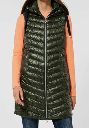 Kvinde iført lang, mørkegrøn quiltet vest uden ærmer med lynlås foran og lommer over en ferskenfarvet langærmet skjorte og blå jeans.