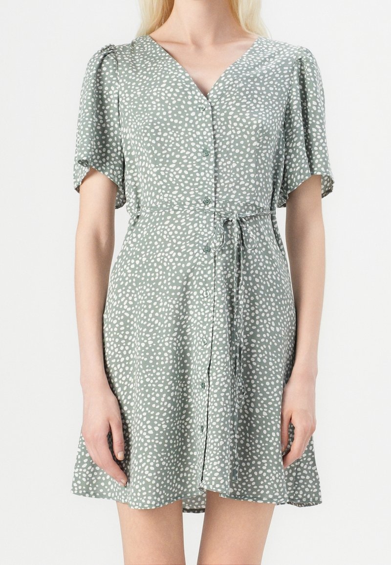 Femme portant une robe courte à manches courtes, vert clair à pois blancs, boutonnée devant, col en V et ceinture nouée à la taille, debout devant un fond uni.