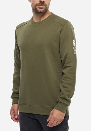Homme portant un sweat-shirt à manches longues vert olive avec des poignets côtelés et des logos sur la manche gauche, associé à un pantalon foncé, debout devant un fond blanc.