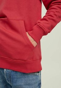 Persona con un sudadera roja, con una mano en el bolsillo canguro frontal, combinada con unos jeans de mezclilla azules, sobre un fondo claro.