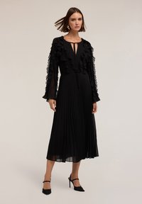Schwarzes, plissiertes Kleid mit langen Rüschensäumen, Schlüsselloch-Ausschnitt und fließendem Stoff. Getragen mit schwarzen High-Heels.