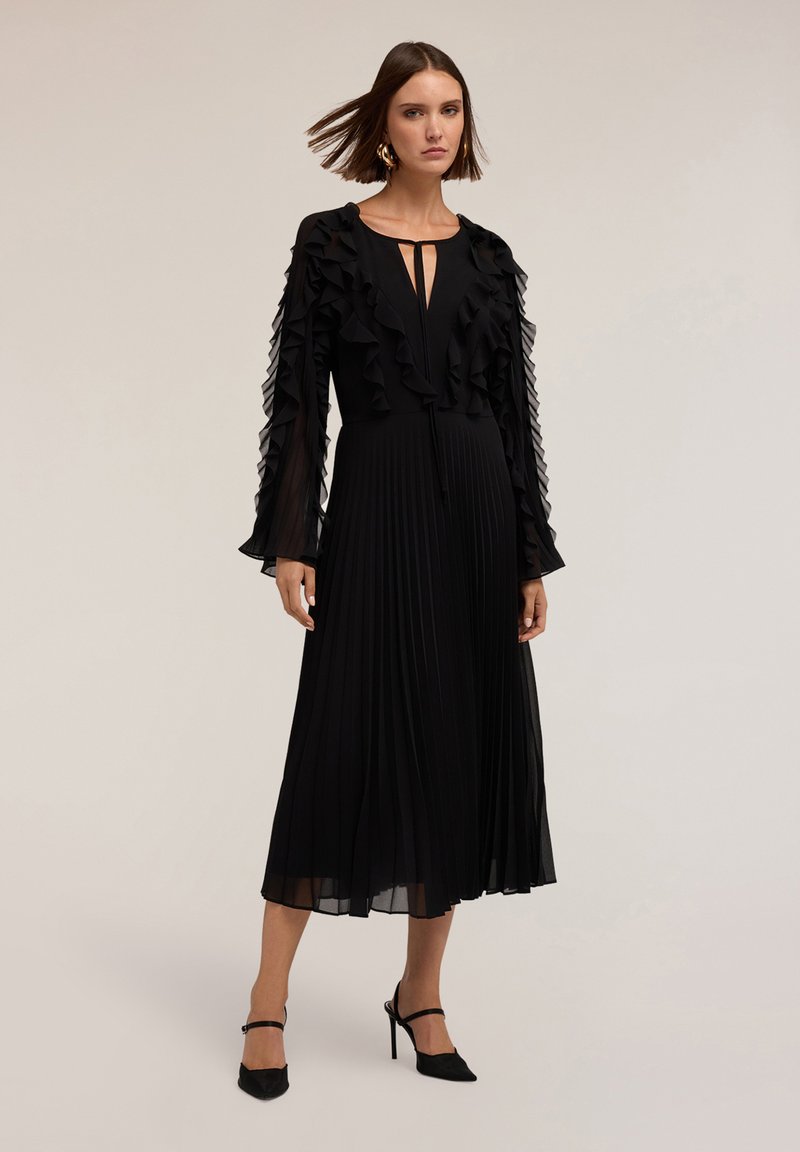 Schwarzes, plissiertes Kleid mit langen Rüschensäumen, Schlüsselloch-Ausschnitt und fließendem Stoff. Getragen mit schwarzen High-Heels.