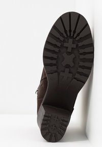 Semelle d'une botte brun foncé avec un motif de crampons profonds comprenant des croix et des formes géométriques, inclinée contre un sol et un mur blancs.