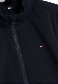 Marinblå vindjacka med hög krage, framdragkedja och liten logotypbård med röd, vit och blå accent. Slät yta.