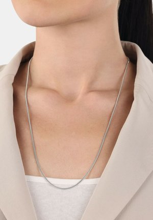 Un collar de cadena de plata reposa sobre una clavícula desnuda, con un diseño suave y elegante. Lleva puesto un top de color claro y un blazer beige.