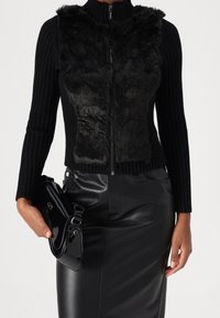 Kvinde iført en sort ribstrikket sweater med lynlås og en blød forside, sort lædernederdel og med en skinnende sort håndtaske i hånden.