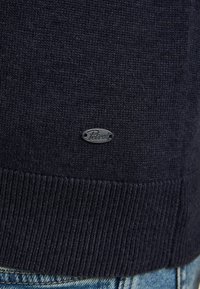 Marineblauwe gebreide trui met een geribbelde zoom en een klein zwart ovaal label met de tekst 'Petrol' nabij de onderrand. Zachte textuur, casual ontwerp.