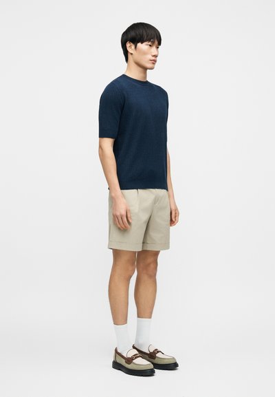 Chemise à manches courtes en maille texturée bleu marine, associée à un short beige. Le mannequin porte des chaussettes blanches et des chaussures grises avec des accents marron.