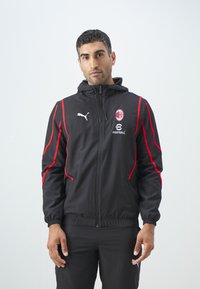 Chaqueta negra con acentos rojos, cierre de cremallera, capucha y logo. Confeccionada en material ligero, diseñada para el fútbol o ropa deportiva.
