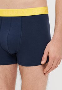 Boxers em algodão azul-marinho com um cós elástico amarelo com a palavra "HAPPY" em relevo. Vista em close-up mostrando o design lateral e frontal.
