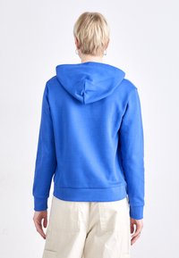 Hanorac albastru cu glugă și siret, cu manșete ribbate, fabricat dintr-un material texturat. Puratat peste o pereche de pantaloni cargo de culoare deschisă.
