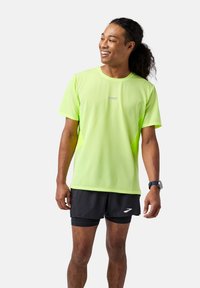 Neon žalia, trumpomis rankovėmis sportinė marškinėlė su tekstūruotu raštu, derinama su juodomis šortais, kurie turi baltą logotipą. Tinkama sporto veikloms.