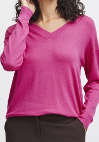 Pull col en V rose en tissu maille douce, avec manches longues et coupe décontractée. L'encolure est ornée de détails côtelés.