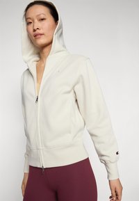 Light beige zip-up hoodie made from soft fabric, featuring a drawstring hood, ribbed cuffs, and a small logo on the chest.