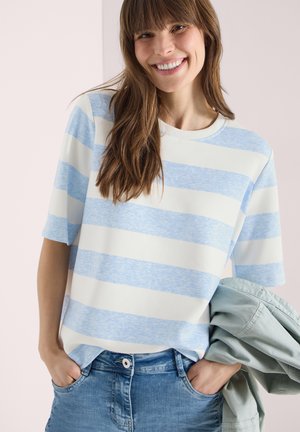 Donna sorridente con capelli lunghi castani, che indossa una maglietta a maniche corte a righe blu e bianche, jeans blu e tiene in mano una giacca leggera.