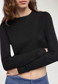 Maglione nero a maglia con scollo rotondo, maniche lunghe e polsini a costine, indossato con pantaloni blu a vita alta. Tessuto liscio.