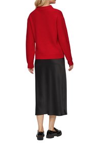 Czerwony sweter z prążkowanej tkaniny z wysokim kołnierzem, noszony na czarnej satynowej spódnicy midi, połączony z czarnymi błyszczącymi butami.