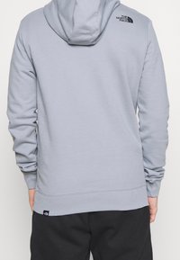 Sudadera gris claro con capucha, con puños y bajo acanalados, y un logo negro en la parte superior de la espalda. Textura suave, diseño casual.
