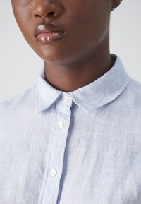 Jasnoniebieska lniana koszula z kołnierzykiem typu button-down, z białymi guzikami, subtelną teksturą oraz małym haftowanym logo na piersi.