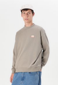 Grå hettegenser med rund hals og ribbestrikkede mansjetter, som har et lite fargerikt merke på brystet. Brukt med lyseblå denimbukser.