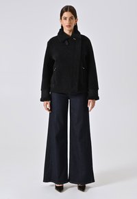 Giacca nera con texture, colletto alto e zip frontale, abbinata a jeans in denim scuro a gamba larga e tacchi a punta neri.