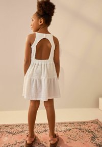 Vit ärmlös klänning med strukturerad tyg, öppen ryggdesign och draperad kjol. Bärs med bruna sandaler på en mönstrad matta.