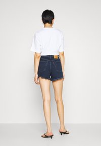 Shorts en denim avec un lavage bleu foncé, ourlet effiloché et poches arrière. Associés à un t-shirt blanc décontracté et des sandales noires.