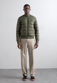Olivengrøn pufferjakke med lynlås foran, ribbede manchetter og kant, parret med beige cargo-bukser og sorte sneakers med orange såler.