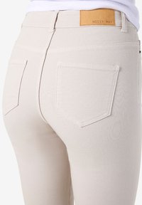 Κοντινό πλάνο μπεζ ψηλόμεσου τζιν με πίσω τσέπες και δερμάτινο ταμπελάκι επωνυμίας "NOISY MAY" που φοριέται με λευκό τοπ.