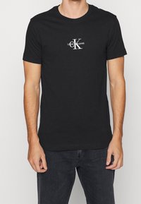 Camiseta de algodón negra de manga corta con un logotipo en blanco "CK" y el texto "CALVIN KLEIN JEANS" impreso en una fuente sencilla.