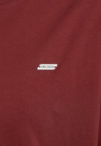 Burgundy tyg med slät yta som har en liten silveretikett märkt "SMILODOX", placerad nära nedre högra hörnet.