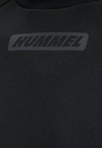 Sort atletisk top med et struktureret stof og mat overflade, der tydeligt viser logoet "HUMMEL" i en mørkegrå, rektangulær label.