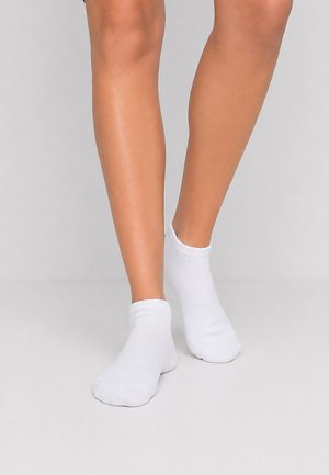 Jambes portant des chaussettes blanches basses unies, debout sur un sol gris clair sur fond gris.