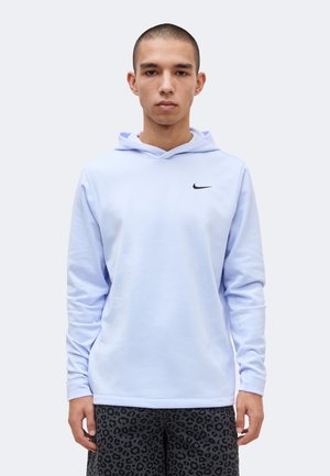 Νέος άνδρας με ανοιχτό μπλε φούτερ Nike και μαύρα σορτς με λεοπάρ μοτίβο, στέκεται μπροστά σε λευκό φόντο.