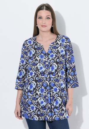 Frau trägt eine schwarz-weiße Tunika mit blauen Blumenmustern und Dreiviertelärmeln, stehend vor einem einfarbigen hellen Hintergrund.